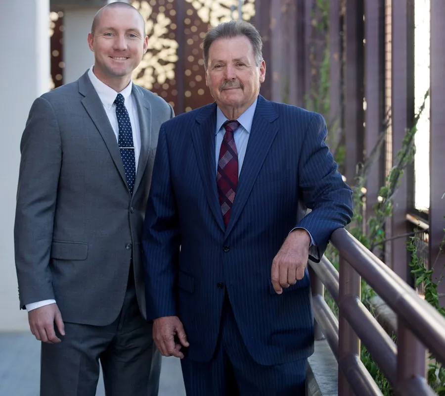 Two professional male attorneys in suits standing on an outdoor walkway, smiling at the camera. One lawyer wears a gray suit; the other wears a navy pinstripe suit.