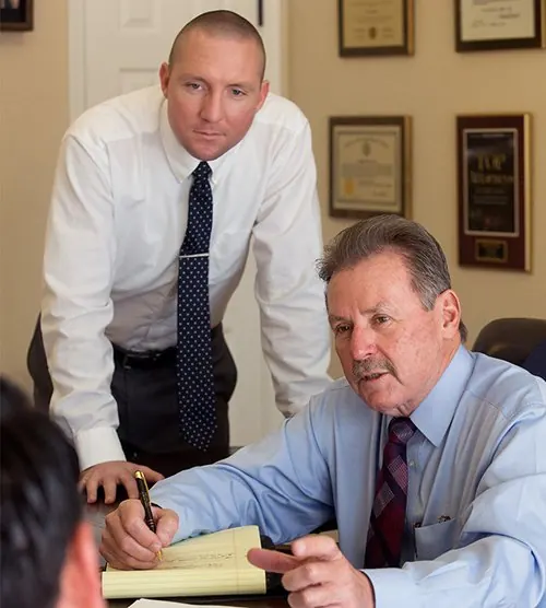 Attorneys Ralls and Wille in their law office conducting a meeting with a client. The seated attorney is writing notes on a legal pad and gesturing, while the other stands behind him observing. Framed legal certificates are visible on the wall.